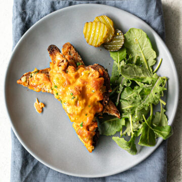 Green salad, sliced pickles and two triangular halves of a salmon melt on a plate that is placed on a cloth napkin.