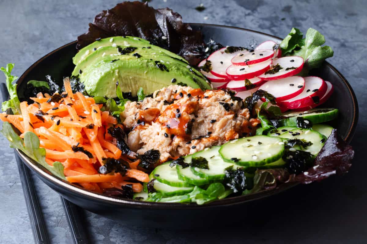 A sushi bowl topped with sliced avocado, radishes, carrot and cucumber and creamy salmon salad.