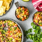 Bowls of colorful chopped veggies and corn alongside avocado halves and tortilla chips.