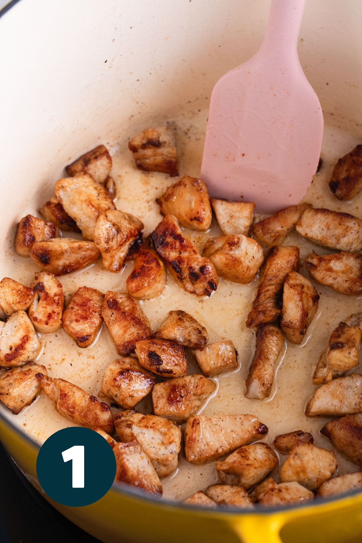 Cubes of browned chicken cooking in an enamelled cast iron pot.