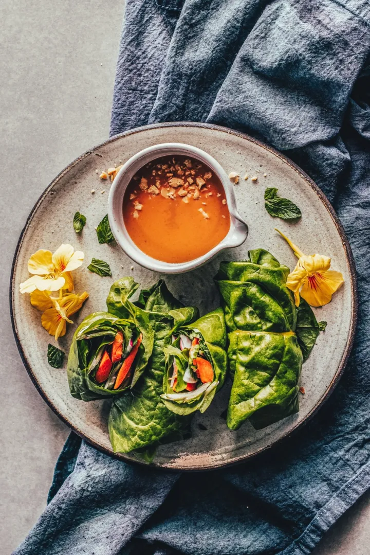 Swiss chard wrapped vegetable rolls on a plate with a bright peanut dipping sauce in a bowl and some nasturtium flowers around.