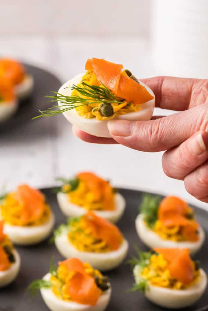 A hand holding a devilled egg topped with lox, capers and a sprig of dill above a platter of devilled eggs.
