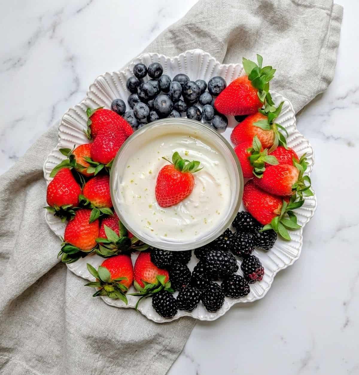 A round fruit platter of strawberries, blueberries and blackberries surrounding a bowl of creamy dip with a strawberry on top.