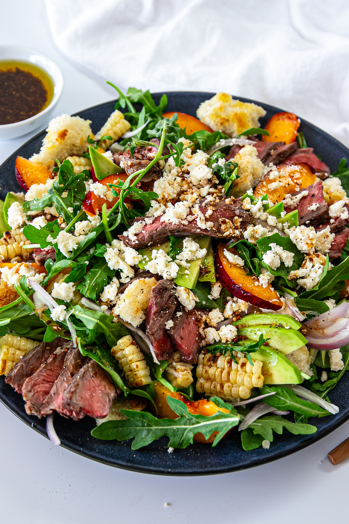 Serving plate of steak and arugula salad with peaches, corn, avocado and a sprinkle of feta over top.