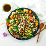 A round serving platter of arugula salad topped with sliced steak, peaches and avocado as well as crumbled feta and grilled corn kernels.