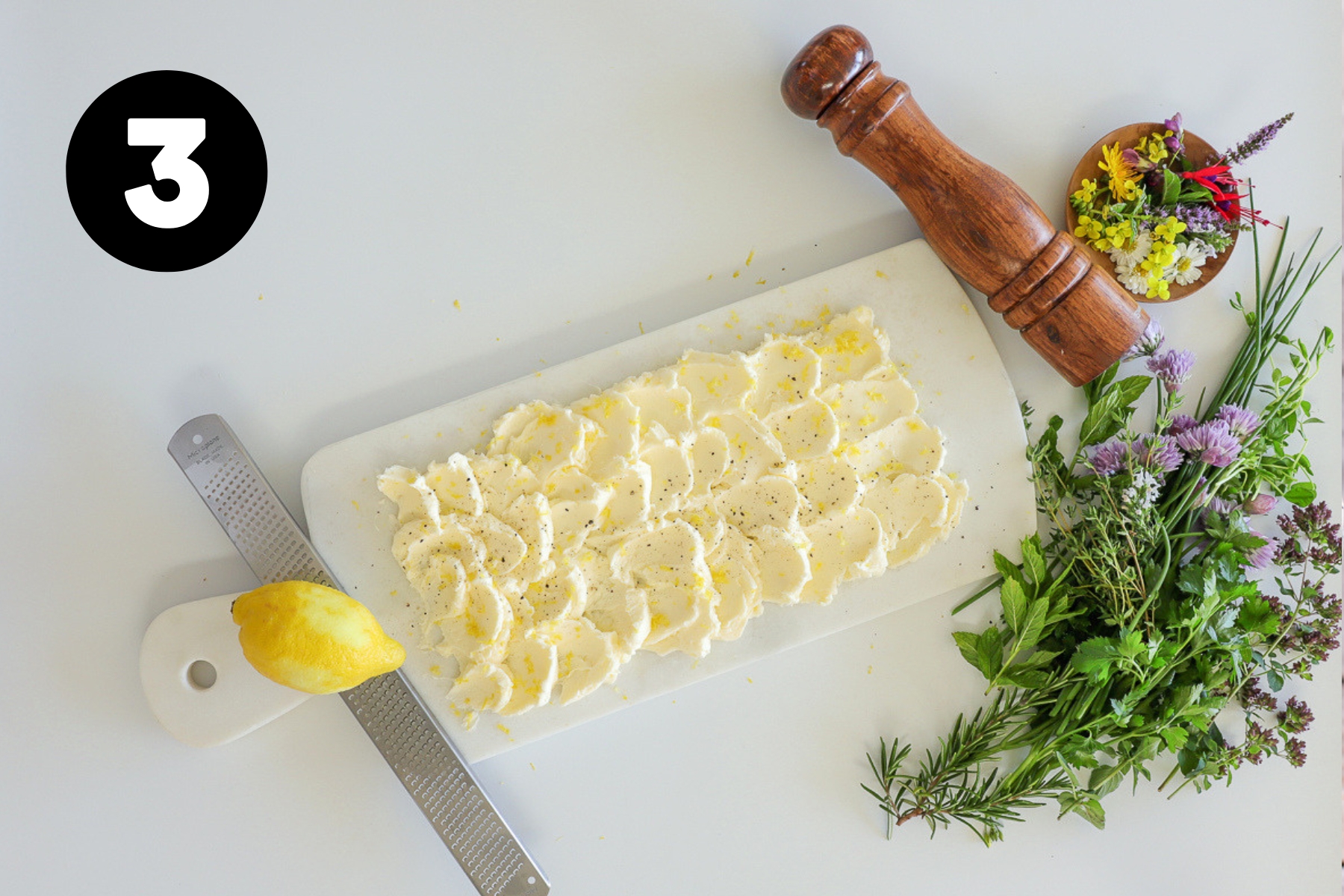 A microplane grater with a lemon sitting on it, lemon zest sprinkled over the butter board and a pepper mill beside the board.