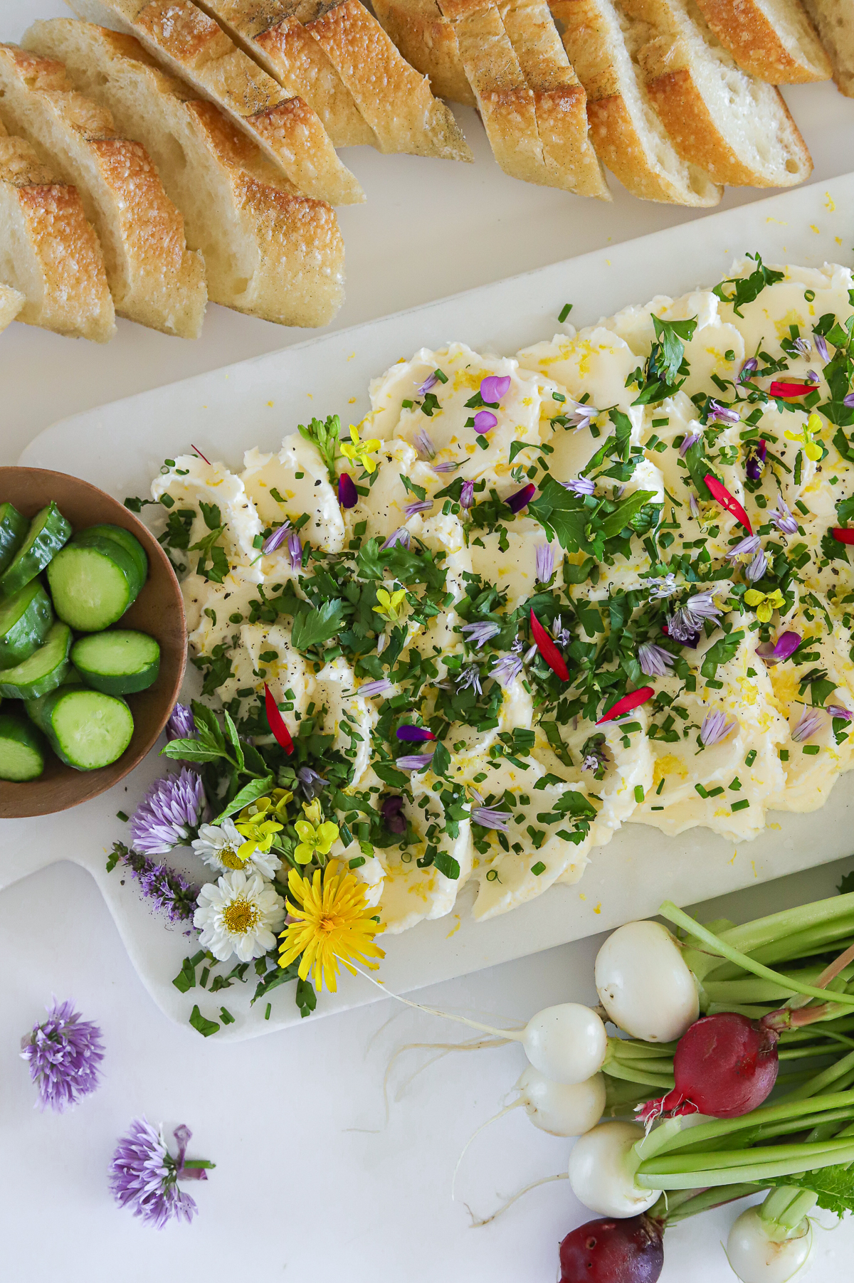 Slice baguette, fresh sliced cucumber, whole radishes and flowers around an herb laden butter board.