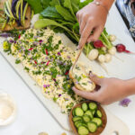 Hands spreading the herb flecked butter onto a piece of bread beside the herb and flower decorated butter board.