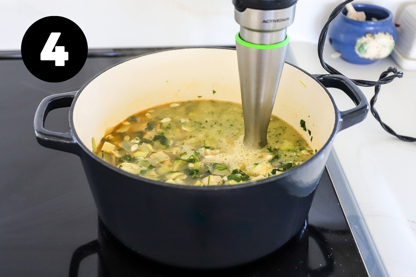 A stick blender is in the pot of soup and half is pureed, while the one side still looks chunky.