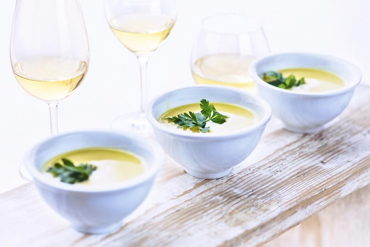 Three bowls of creamy soup garnished with parsley on a wooden table with 3 glasses of white wine behind,