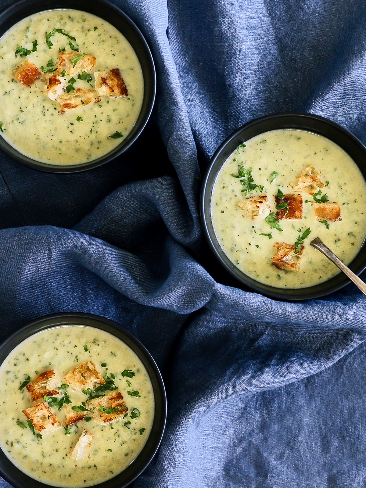 3 bowls of light, creamy soup topped with croutons on a navy blue linen tablecloth.